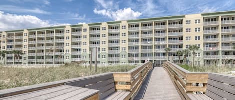 Una pasarela de madera que conduce a un edificio de varios pisos con balcones, con vista a una playa.