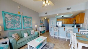 A spacious living room with a teal sofa, a coffee table, and a kitchen area with wooden cabinets and a white countertop. - Gorgeous beachfront condo w/ocean views, shared pool, and beach access (Fort Walton Beach)