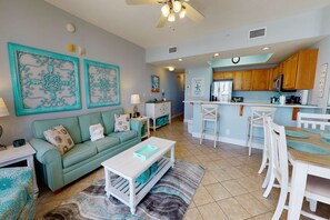 A spacious living room with a teal sofa, a coffee table, and a kitchen area with wooden cabinets and a white countertop. - Gorgeous beachfront condo w/ocean views, shared pool, and beach access (Fort Walton Beach)