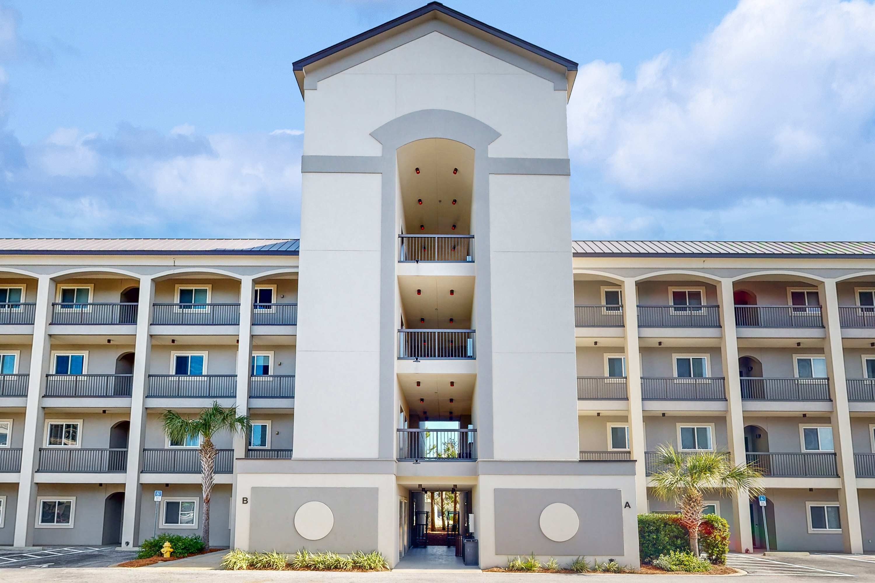 A multi-story hotel with a central entrance, balconies on each floor, and a parking lot in front.