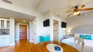 A room with a wooden dining table, a bed with a white headboard, a ceiling fan, and a kitchen area with cabinets and a countertop. - Fourth-floor studio with shared pool, fast WiFi, washer&dryer, & central AC (Miramar Beach)