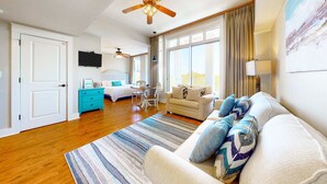 A spacious living area with a large sofa, a coffee table, and a ceiling fan. There is a television mounted on the wall and a window with curtains. - Fourth-floor studio with shared pool, fast WiFi, washer&dryer, & central AC (Miramar Beach)