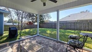 Eine überdachte Terrasse mit Holztisch und -stühlen, einem Deckenventilator und Blick auf einen Garten mit Holzzaun und Bäumen.