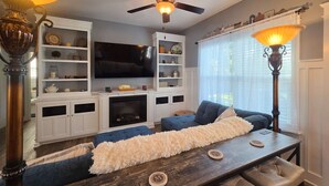 Ein Wohnzimmer mit Kamin, einem an der Wand befestigten Fernseher, einem Deckenventilator und einem Esstisch mit Stühlen.