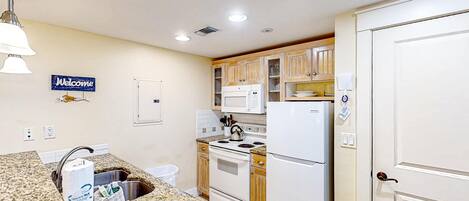 Una cocina con encimeras de granito, estufa, microondas y refrigerador. Hay un fregadero con lavavajilla y un cartel de bienvenida en la pared.