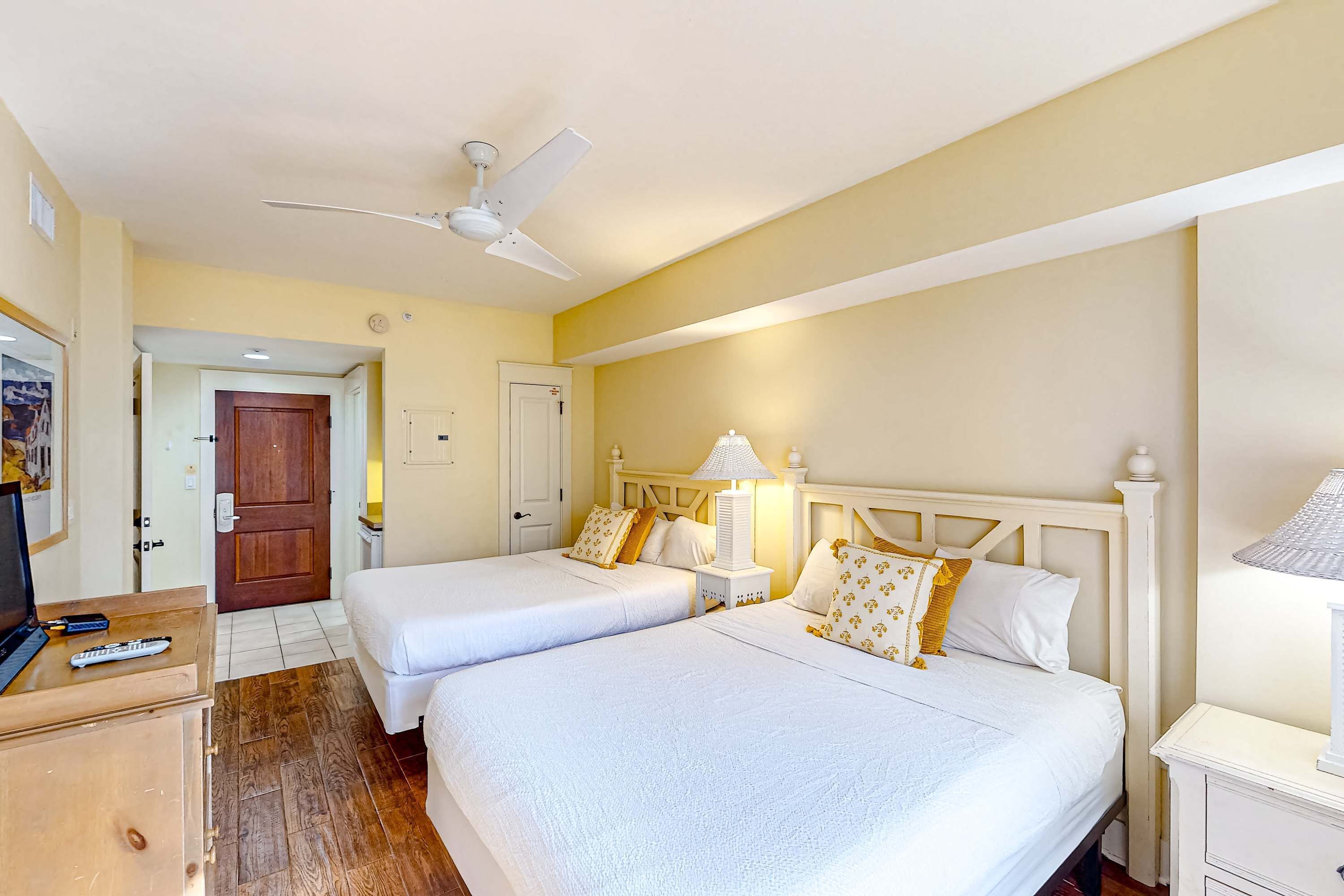 A hotel room with two beds, a desk, a ceiling fan, and a door leading to another room.