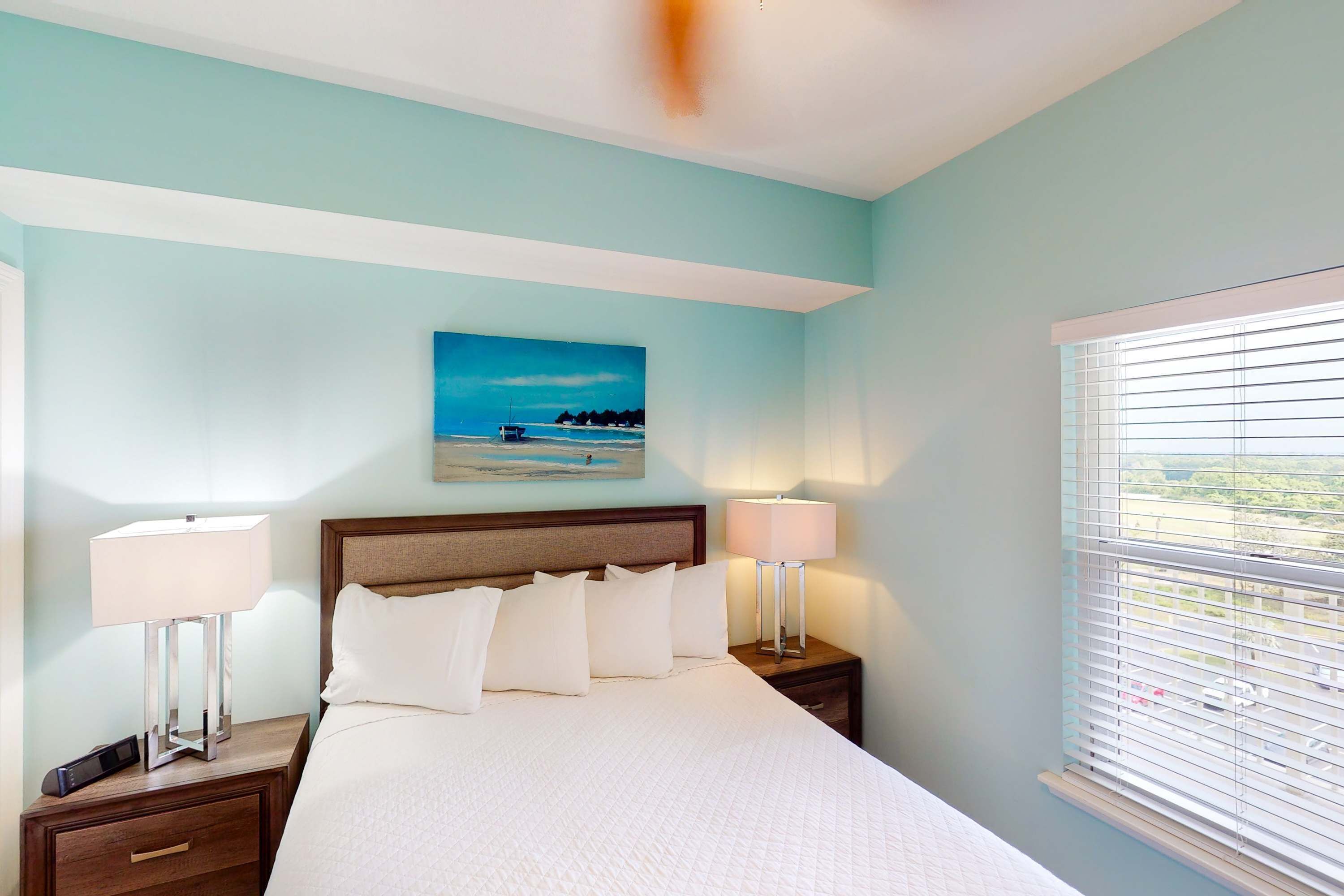 A hotel room with a large bed, two bedside tables with lamps, a painting of a beach scene, and a window with blinds.