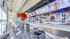 A modern bar area with a long counter, bar stools, and a large screen displaying a person. - Seventh Floor Condo with Beach View Pool Gym and Private Balcony (Miramar Beach)