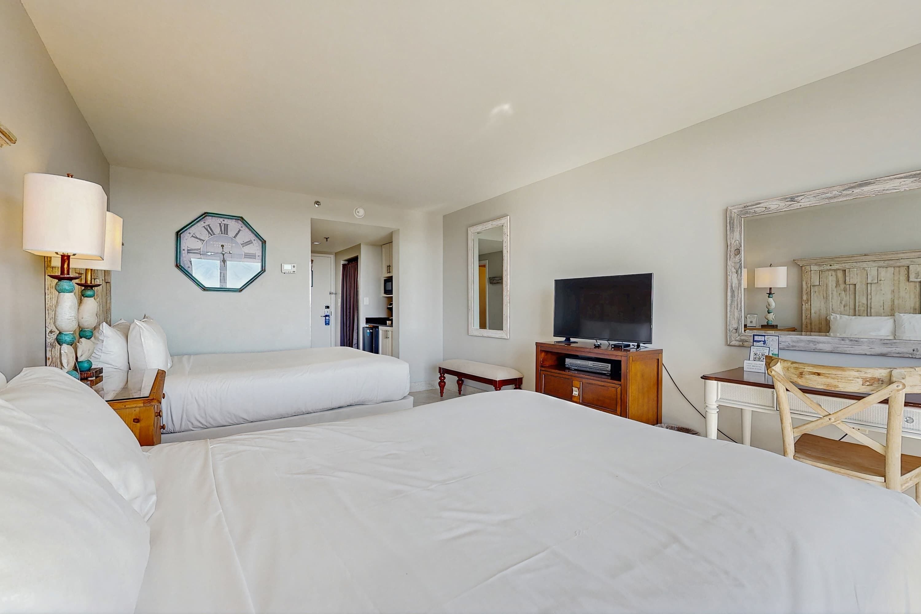 Habitación de hotel con dos camas, televisión, escritorio, silla, reloj y espejo.