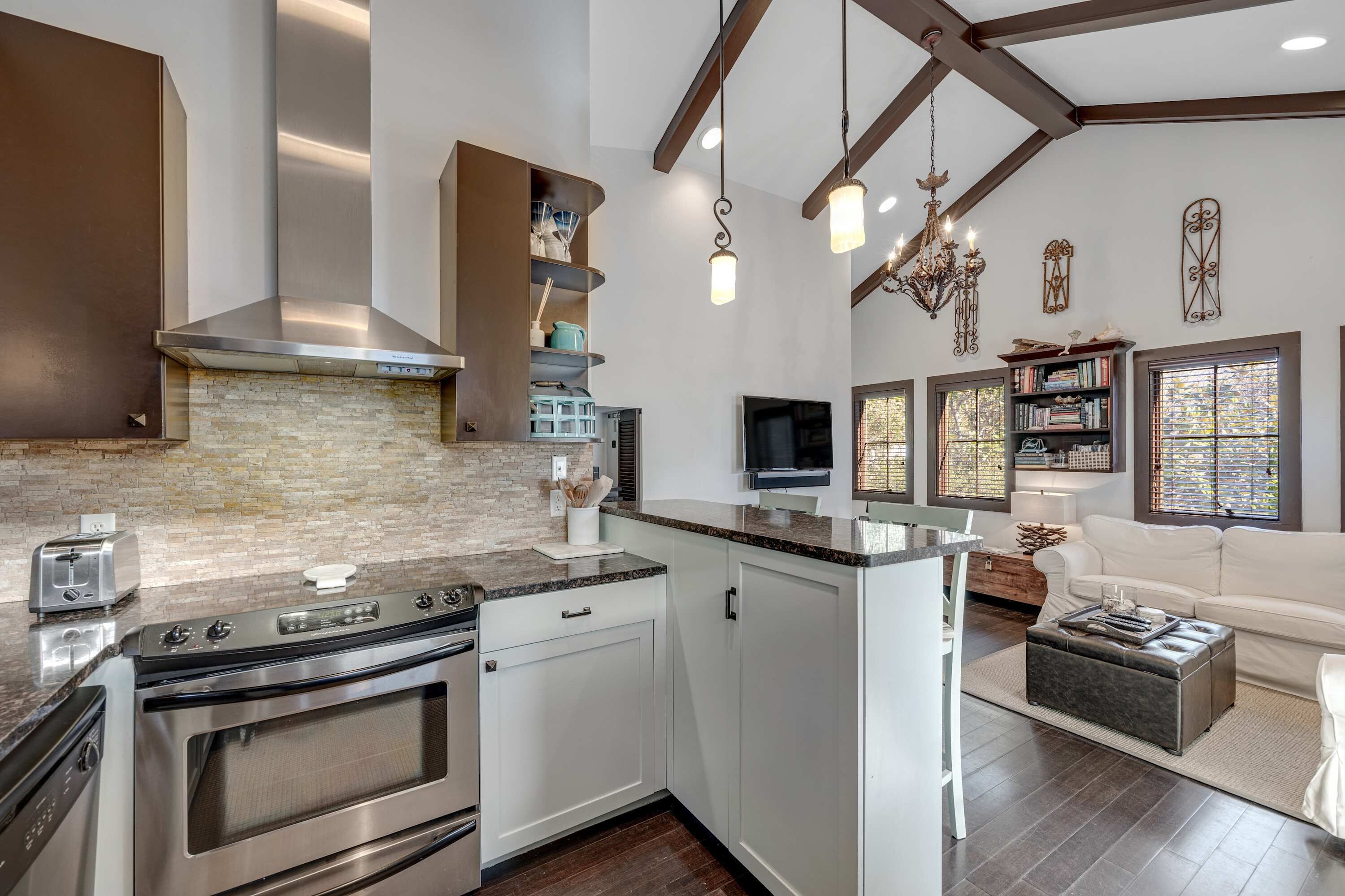 A modern kitchen with a stainless steel range hood, a built-in oven, and a countertop with a toaster. There is a white kitchen island with a sink and a chandelier above.