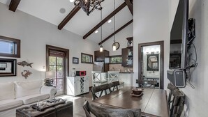 Une cuisine moderne avec des poutres en bois, un coin repas avec une table et des chaises en bois, et un salon avec un canapé blanc et un pouf en cuir.