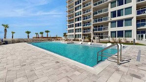 A swimming pool with a herringbone tile pattern, a metal pool ladder, and a multi-story building with balconies in the background. - Beautiful beachfront condo with pool, beach tram, tennis, golf, & on-site dining (Miramar Beach)