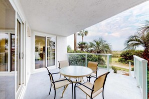 A balcony with a round table and chairs, glass doors, and a view of palm trees and greenery. - Beautiful beachfront condo with pool, beach tram, tennis, golf, & on-site dining (Miramar Beach)