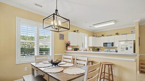 Une cuisine avec des meubles blancs, une table à manger en bois avec des sets de table et un luminaire suspendu.