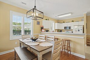 A kitchen with white cabinets, a wooden dining table with placemats, and a hanging light fixture. - Willow Bend 214: 1st Floor End Unit in Barefoot! New On Program! (North Myrtle Beach)