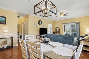 A living room with a dining table, chairs, a sofa, a TV, and a ceiling fan. - Willow Bend 214: 1st Floor End Unit in Barefoot! New On Program! (North Myrtle Beach)