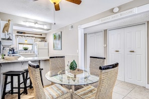 A dining area with a glass table surrounded by wicker chairs, a ceiling fan, and a kitchen in the background. - Convenient condo near beach with pool, balcony, & W/D - snowbird-friendly (Pensacola Beach)