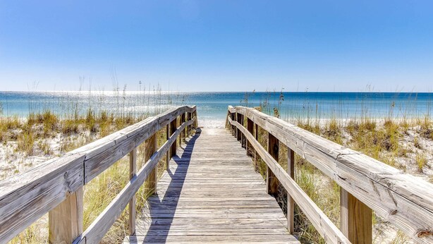 Ein hölzerner Steg führt zu einem Strand bei blauem Himmel.