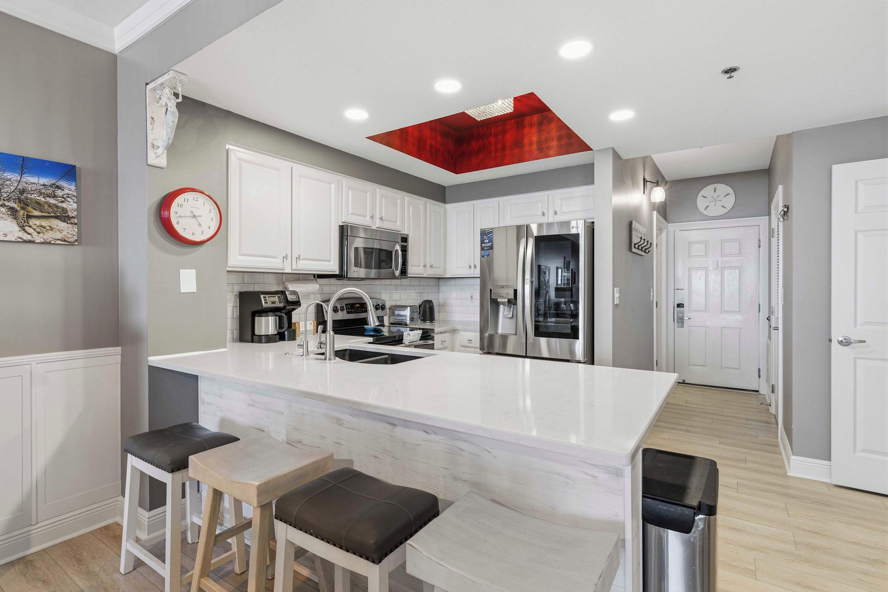 Cocina moderna con armarios blancos, electrodomésticos de acero inoxidable, una isla de mármol con taburetes y un elemento decorativo rojo en el techo.