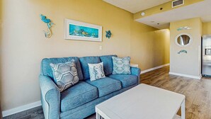 A living room with a blue sofa, white coffee table, and a framed painting on the wall. - Palm Court Yacht Club 312A - Third-floor cozy coastal studio with soundviews, WiFi, and shared pool & hot tub (Fort Walton Beach)