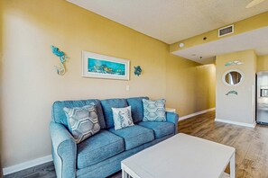 A living room with a blue sofa, white coffee table, and a framed painting on the wall. - Palm Court Yacht Club 312A - Third-floor cozy coastal studio with soundviews, WiFi, and shared pool & hot tub (Fort Walton Beach)
