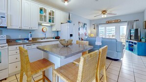 Una cocina con armarios blancos, una isla central con sillas, un ventilador de techo y un letrero que dice 'BEACH HOUSE'.