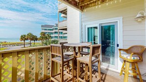Una terraza de madera con una mesa y sillas, con vista a una playa y un edificio con balcones.
