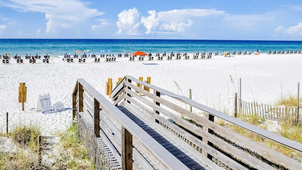 Ein Strand mit einer Holzpromenade, die zum Ufer führt, weißem Sand und blauem Himmel.