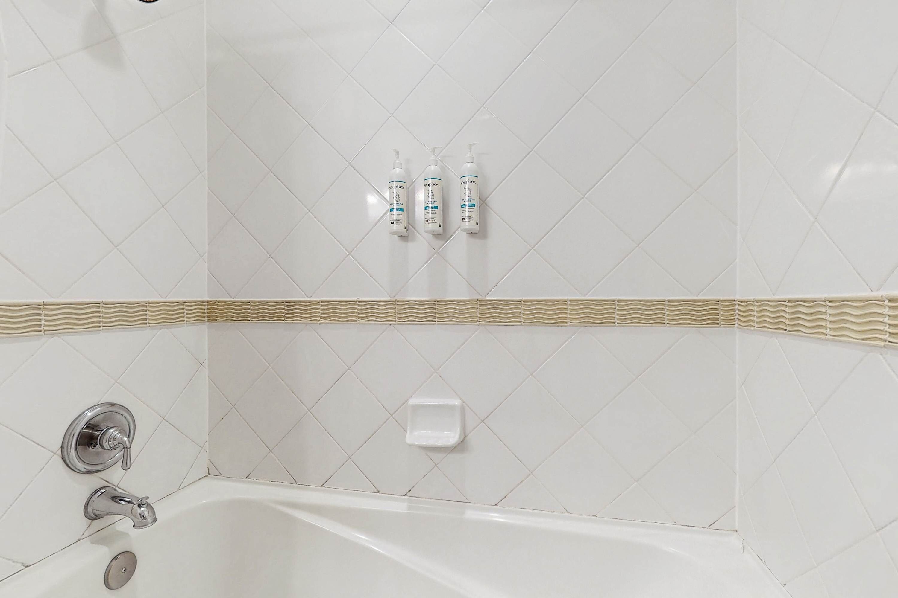 Une salle de bain avec un mur recouvert de carreaux blancs, une baignoire et trois bouteilles de shampoing.