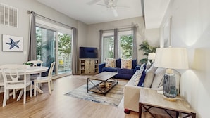 Ein großzügiges Wohnzimmer mit Sofa, Couchtisch und Fernseher. Es gibt einen Essbereich mit Tisch und Stühlen. Das Zimmer verfügt über große Fenster und einen Deckenventilator.