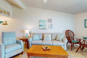A living room with a light blue armchair, a grey sofa with a 'Life is Simply Better at the Beach' pillow, a wooden coffee table, and a wooden dining table with a chair. - Comfy Coastal Condo w/ Gorgeous Bay Views & Nearby Beach Access (Pensacola Beach)