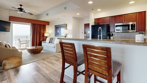 A modern living room with a sofa, coffee table, and wooden chairs. A kitchen area with wooden cabinets and a microwave is visible. - Resort condo w/gulf view, corner balcony & shared pool/hot tub/beach access (Miramar Beach)