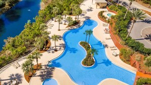 A large, winding pool surrounded by palm trees and lounge chairs, with a small island in the center. - Resort condo w/gulf view, corner balcony & shared pool/hot tub/beach access (Miramar Beach)
