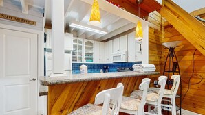 Une cuisine avec des meubles blancs, un pare-feu en carreaux bleus et un plafond en bois. On y trouve des chaises blanches avec des coussins à motifs et un tabouret de bar.