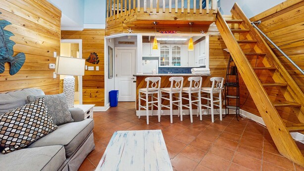 Un vaste espace de vie agrémenté d’un escalier en bois, une cuisine avec des tabourets de bar et un coin salon confortable avec un canapé et des coussins à motifs.
