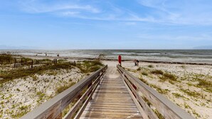 Ein hölzerner Steg führt zu einem Strand mit Dünen und wolkenlosem Himmel.