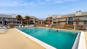 Ein rechteckiger Pool mit klarem, blauem Wasser, umgeben von einer Sandterrasse. Liegestühle und Sonnenschirme säumen den Poolbereich.