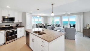 A modern kitchen with stainless steel appliances, a granite countertop, and a large island with a sink. There is a dining area with a view of the ocean and a ceiling fan. - Blu 301 - Third-floor, Gulf-view condo with pool and hot tub access, central AC, & W&D (Fort Walton Beach)