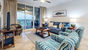 Ein Wohnzimmer mit einer Couch, Sesseln, einem Couchtisch, einem Fernseher auf einem Standfuß und einem Deckenventilator.