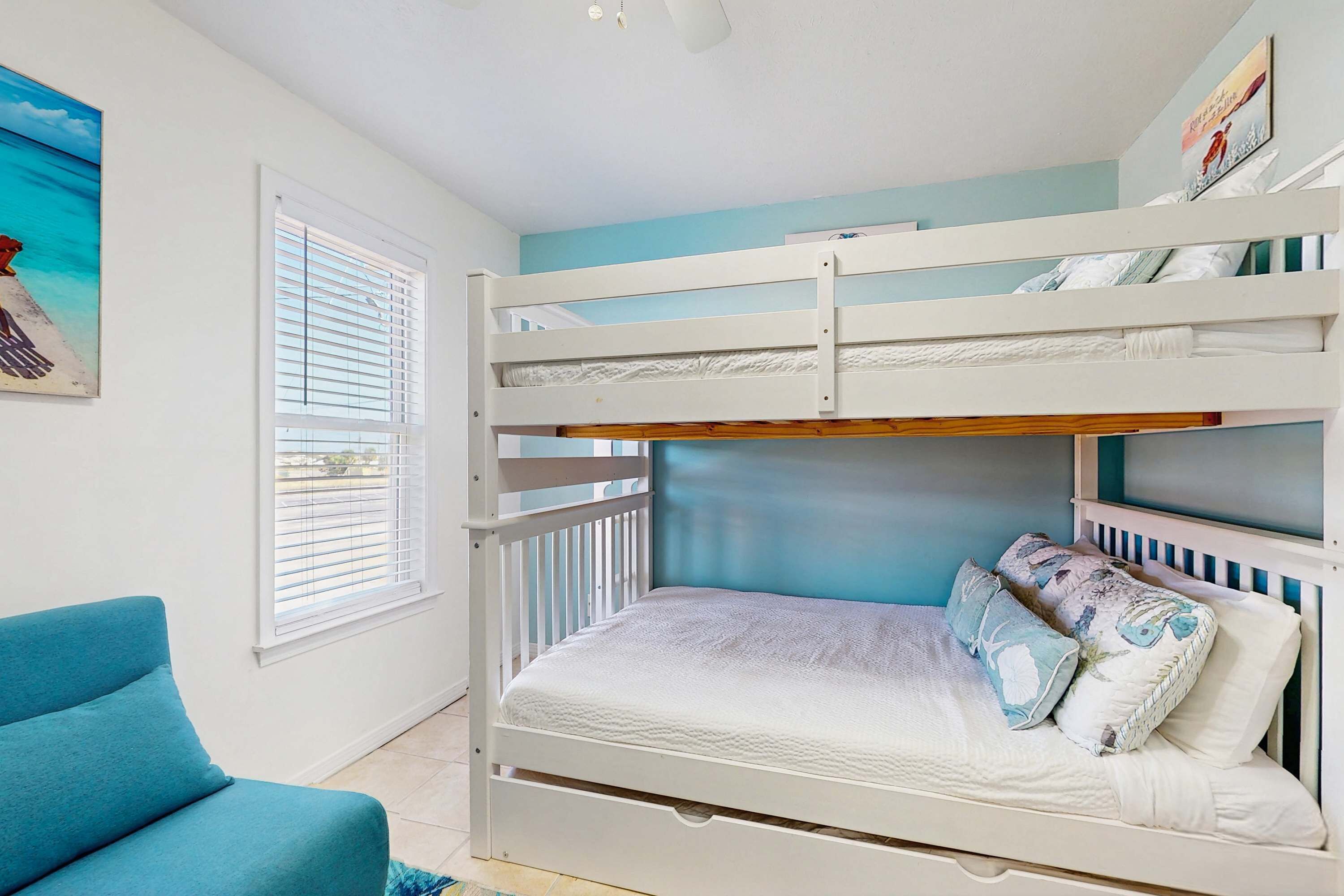 A bunk bed with a lower bunk and a top bunk, a window with blinds, a blue chair, and a painting on the wall.