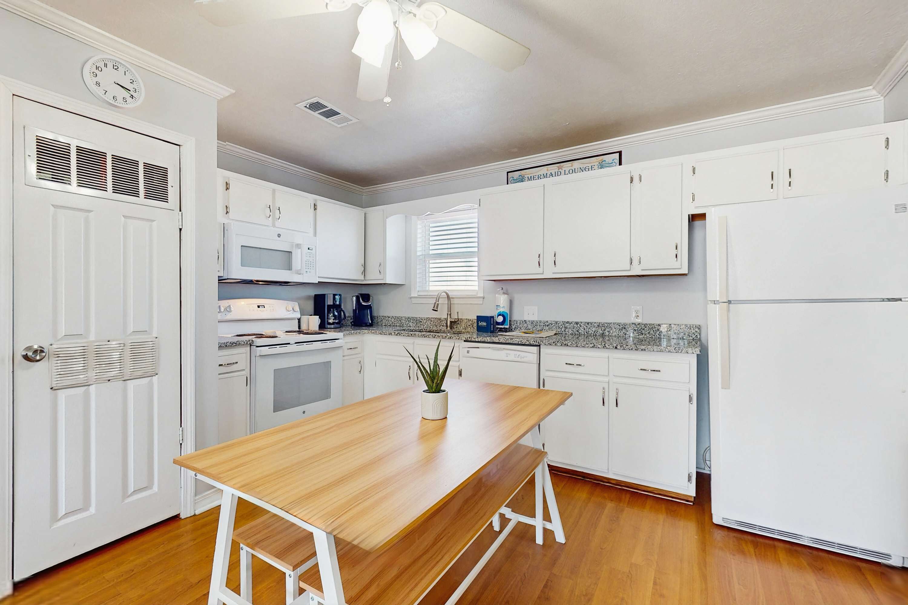 A kitchen with white cabinets, a wooden table, a ceiling fan, and a clock on the wall.