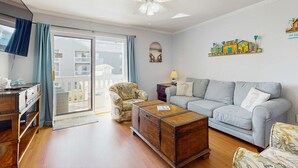 A living room with a sofa, armchairs, a wooden coffee table, a ceiling fan, and a view through a sliding glass door to a balcony. - Cozy sound side condo with shared pool, Close to shopping and beach (Fort Walton Beach)