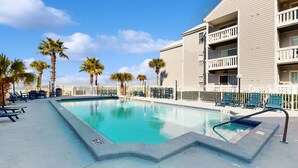 A swimming pool with lounge chairs and palm trees, surrounded by a fence and a building with balconies. - Cozy sound side condo with shared pool, Close to shopping and beach (Fort Walton Beach)