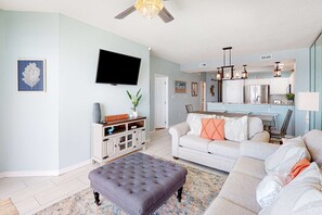 A spacious living room with a grey tufted ottoman, a white sofa, and a kitchen area with white cabinets and a stainless steel refrigerator. - Gulf Dunes 512 - Beachfront condo with gulf views, pool, & balcony - near dining,attractions (Fort Walton Beach)