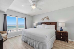 A bedroom with a large bed, a ceiling fan, a nightstand with lamps, a window with curtains, and a view of the beach. - Lovely and Convenient Home In Beachfront Complex w Shared Pool, WiFi, AC, & W&D (Fort Walton Beach)
