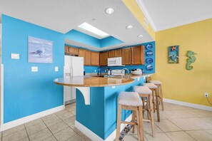 A kitchen with a blue accent wall, wooden cabinets, a white refrigerator, a microwave, and a bar stool seating area. - Beach Chic Condo On Okaloosa Island Gulfside Pool & Exercise Room (Fort Walton Beach)