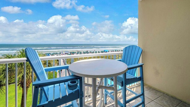 Un balcón con sillas y una mesa azules, con vistas a una playa con sombrillas y gente.