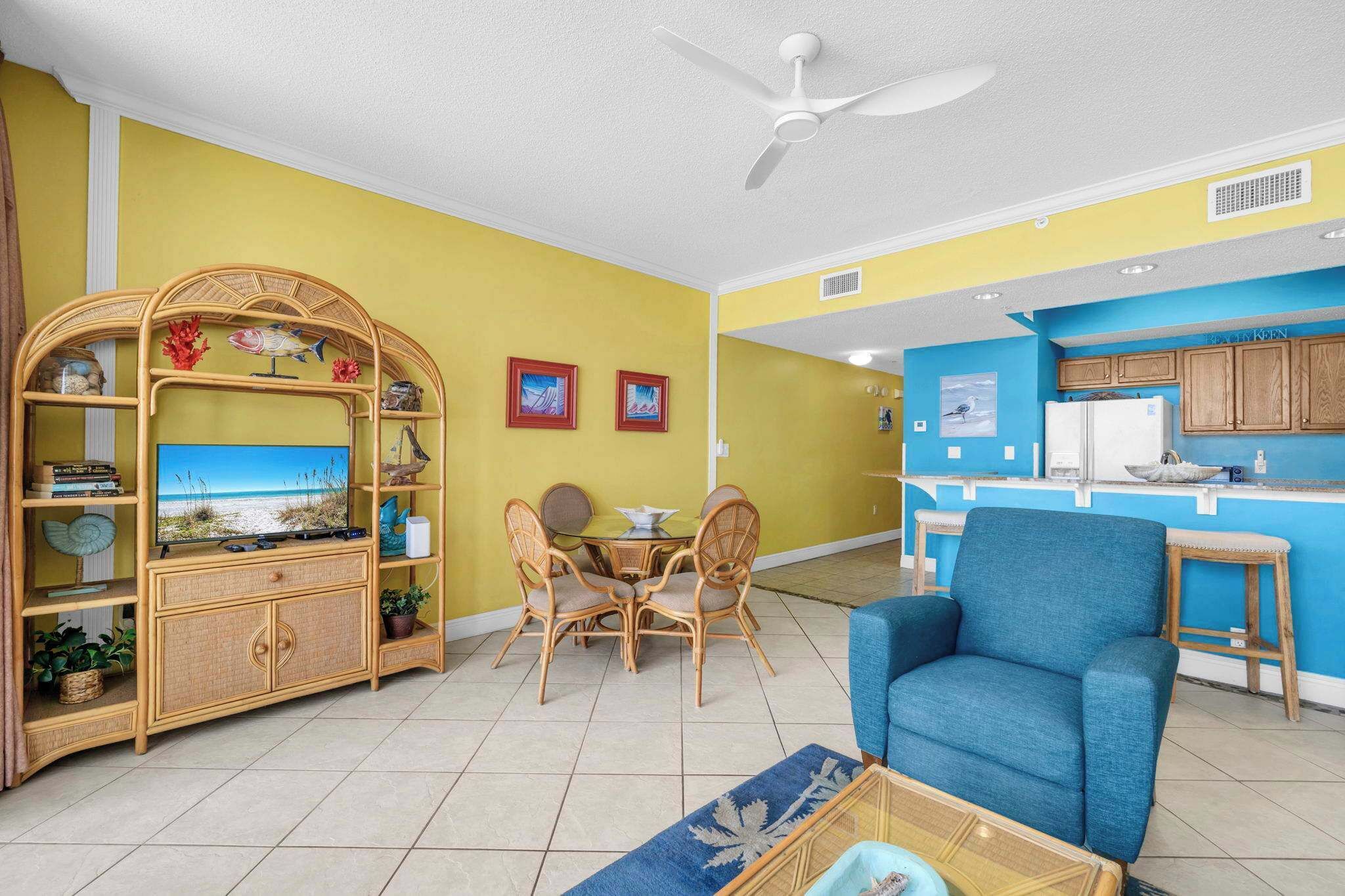 A living room with a dining area, a blue armchair, a ceiling fan, and a kitchen area with wooden cabinets.