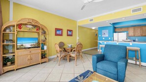 Una sala de estar con comedor, un sillón azul, un ventilador de techo y una zona de cocina con armarios de madera.