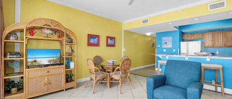 A living room with a dining area, a blue armchair, a ceiling fan, and a kitchen area with wooden cabinets.